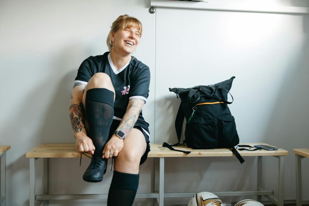Lees alles over voetbal 6 A female soccer player laughing while putting on boots in a locker room.